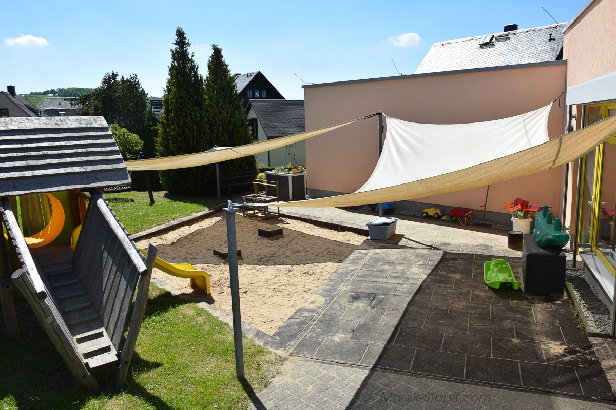 Außenspielbereich der Kindertagesstätte „Regenbogen“ mit Sandkasten, Sonnensegeln, Holzspielhaus mit Rutsche und Spielfahrzeugen