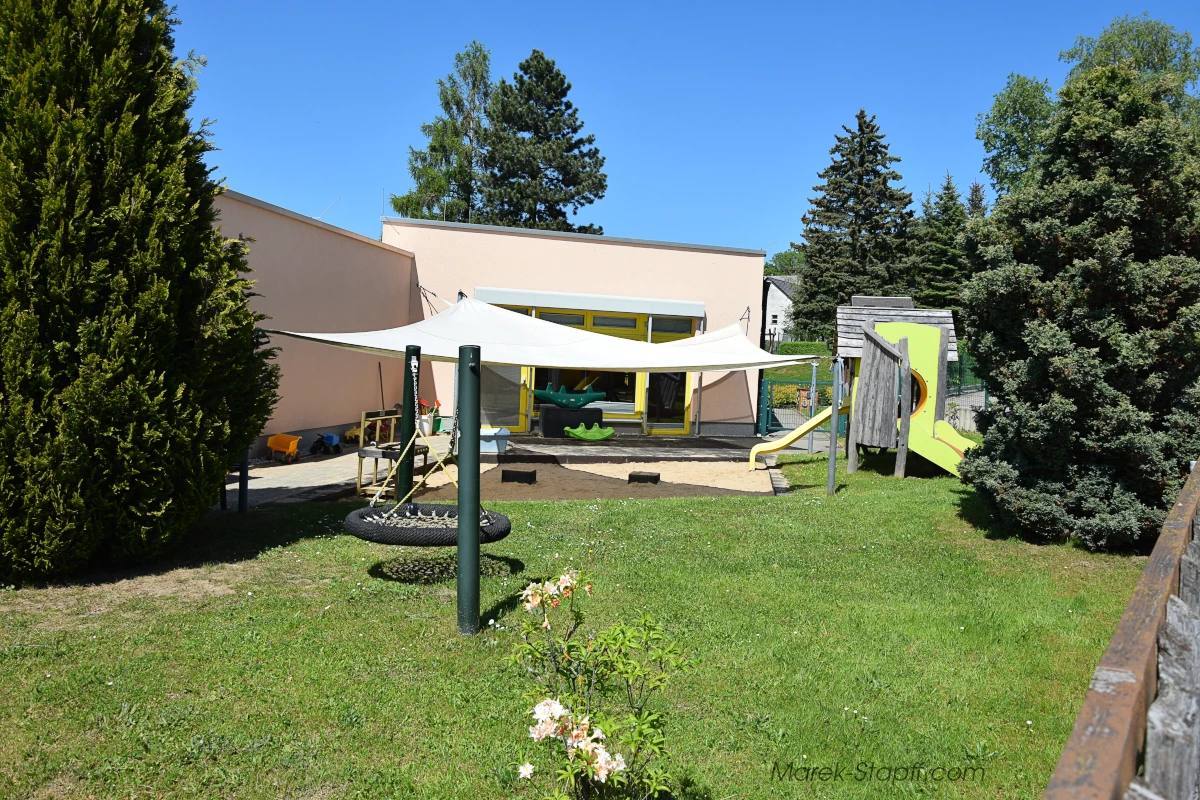 Spielwiese der Kindertagesstätte „Regenbogen“ mit Nestschaukel, Klettergerät und Sonnensegel vor dem Gebäude