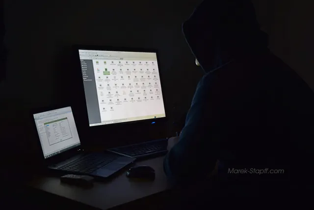 Eine Person mit Kapuze sitzt im Dunkeln vor einem Laptop und einem großen Monitor; nur die Bildschirme leuchten.